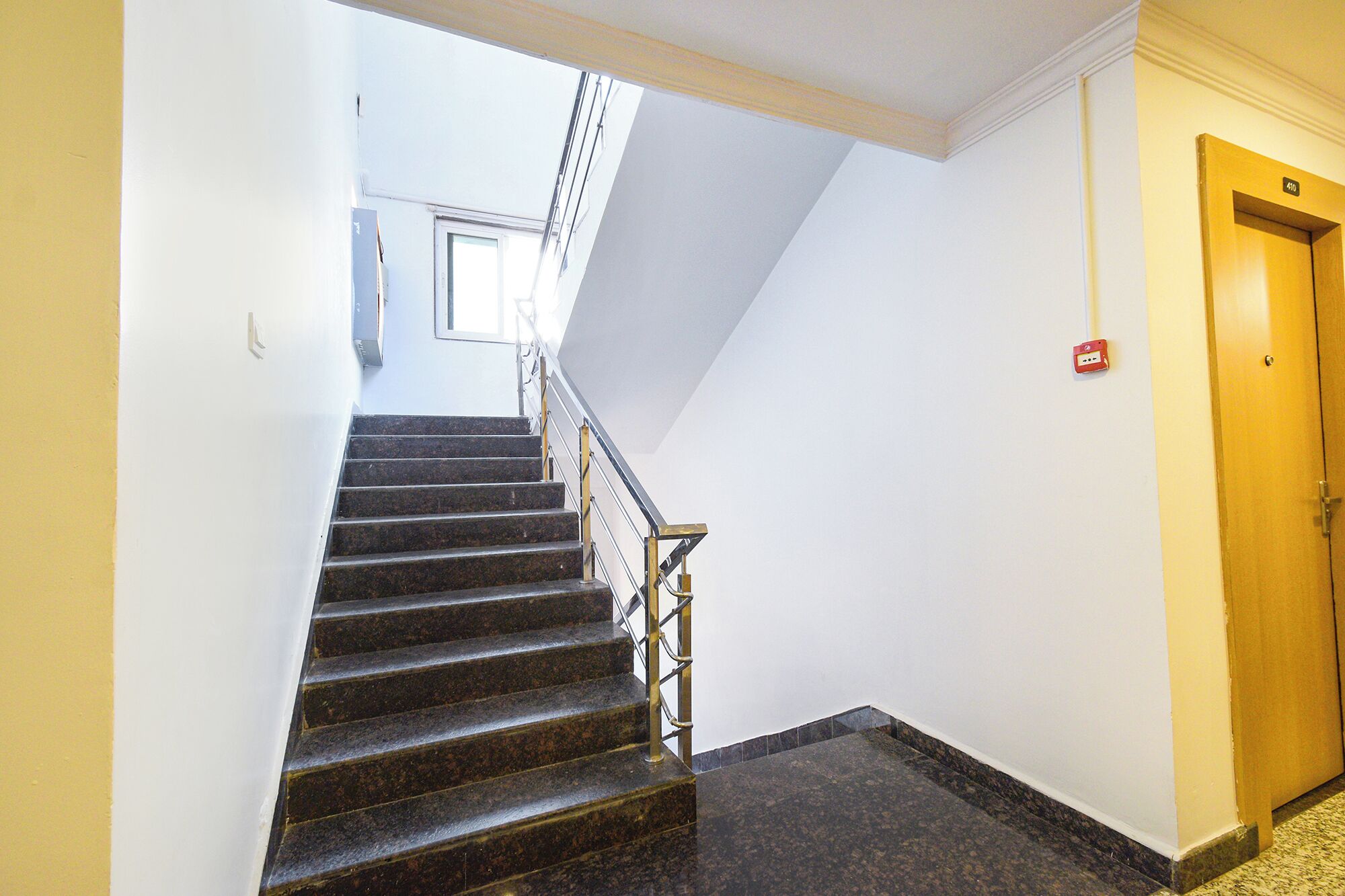 Un escalier avec une rampe métallique menant à un palier, une porte avec une plaque de numéro et une prise électrique fixée au mur.