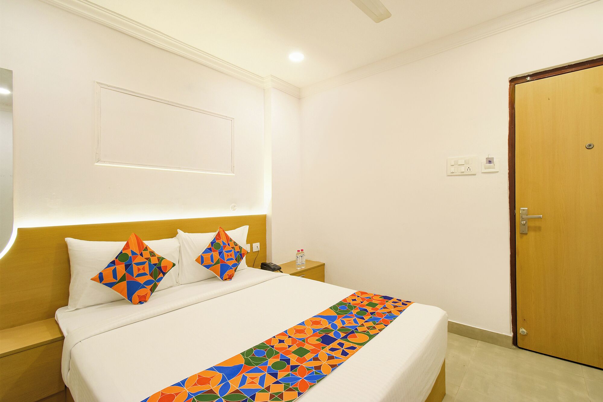 A hotel room with a bed, a nightstand, a bedside table, a wooden door, and a wall-mounted light fixture.