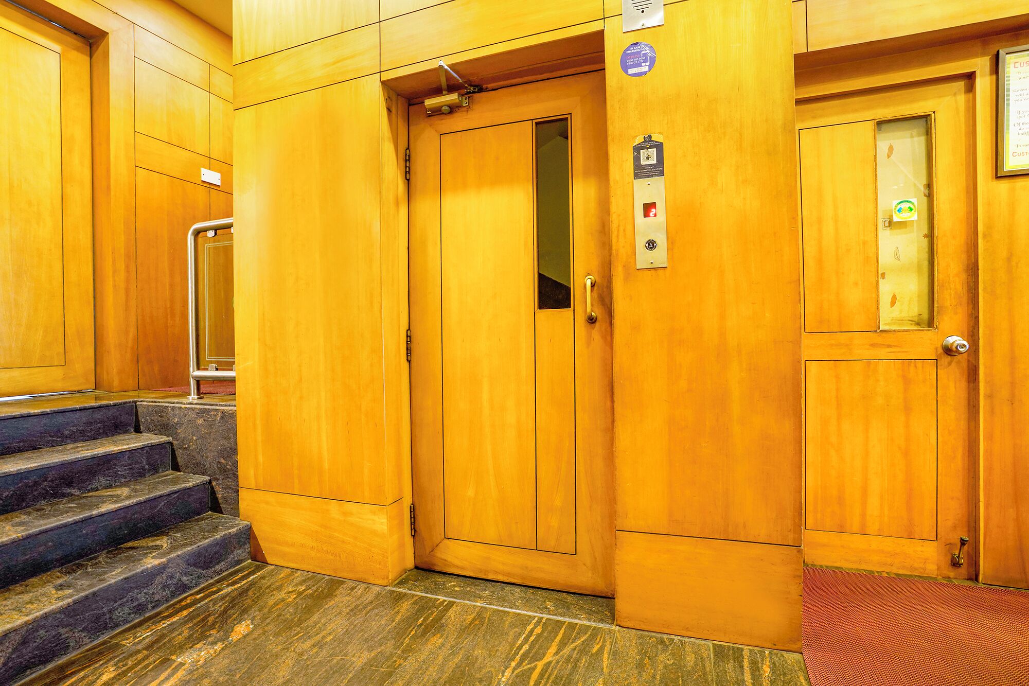 A wooden elevator with a handrail, a control panel, and a red carpet at the bottom.