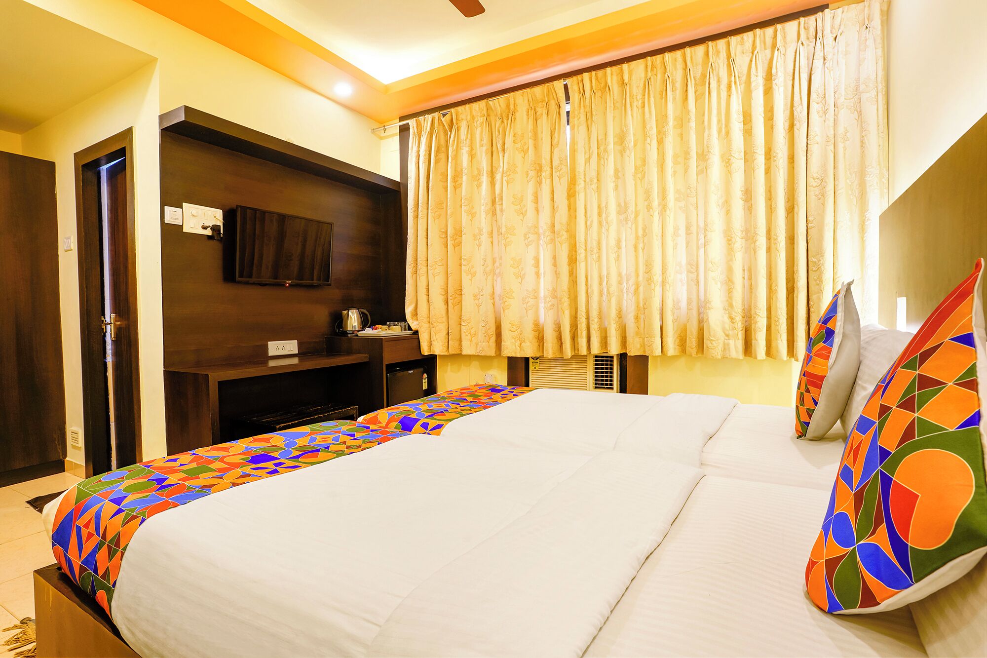A hotel room with a large bed, a TV on a wooden stand, a small table with a kettle, and a window with curtains.