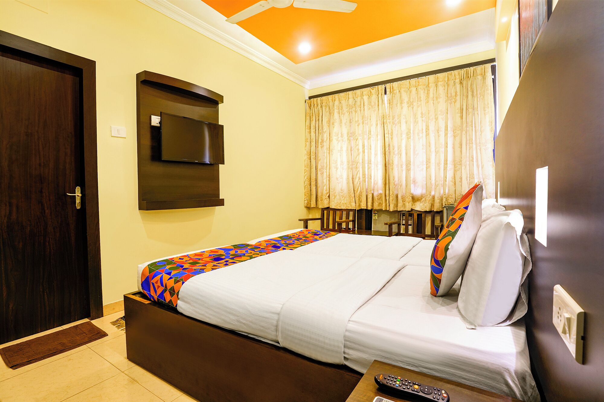 Una habitación de hotel con cama, televisión montada en la pared, ventana con cortinas y ventilador de techo.