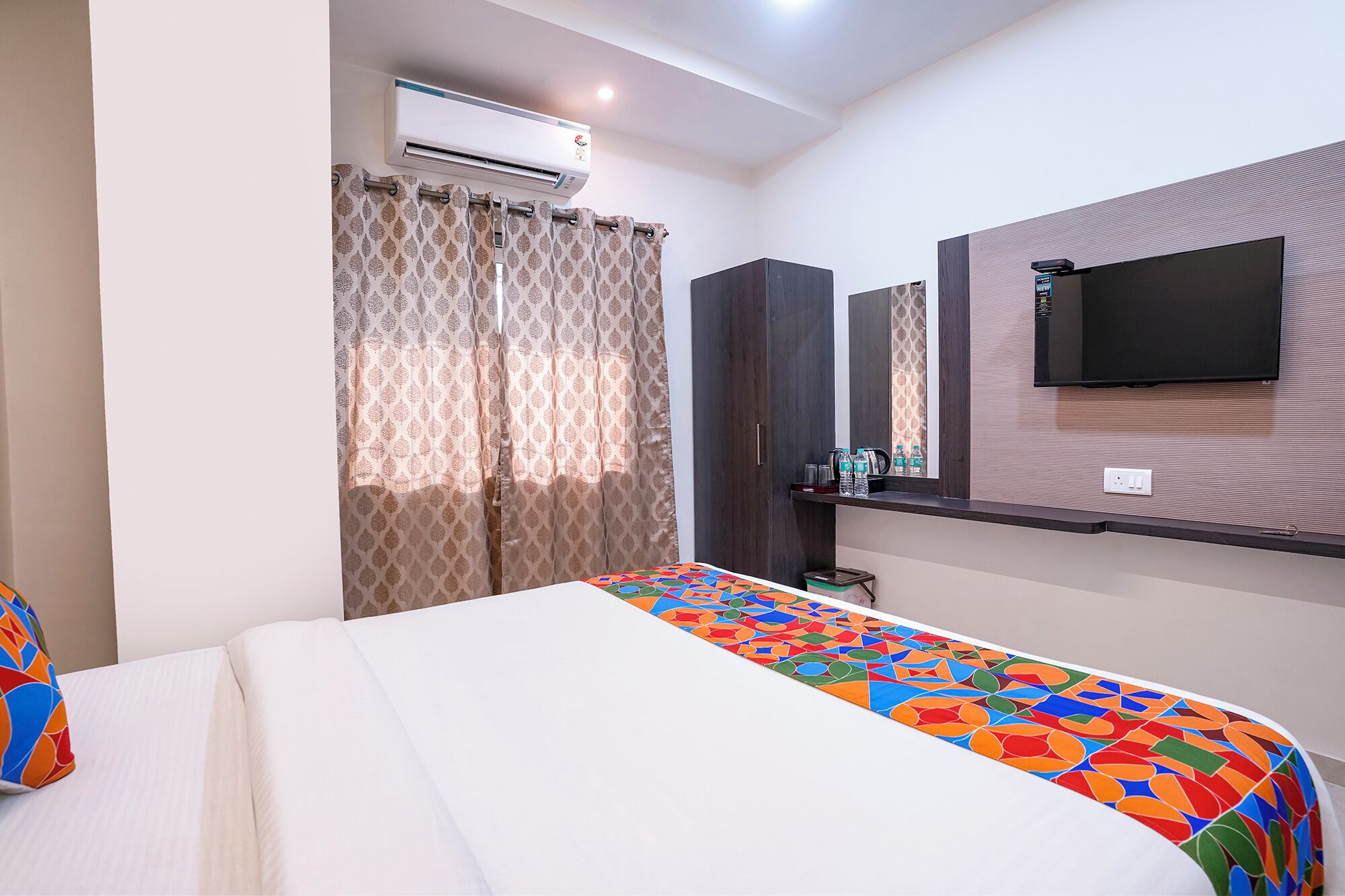 Une chambre d’hôtel avec un lit, un téléviseur fixé au mur, un appareil de climatisation et une fenêtre avec des rideaux à motifs.