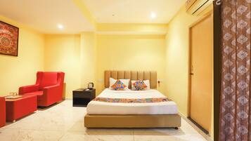 A hotel room with a large bed, a red armchair, a nightstand with a kettle, and a large mirror.