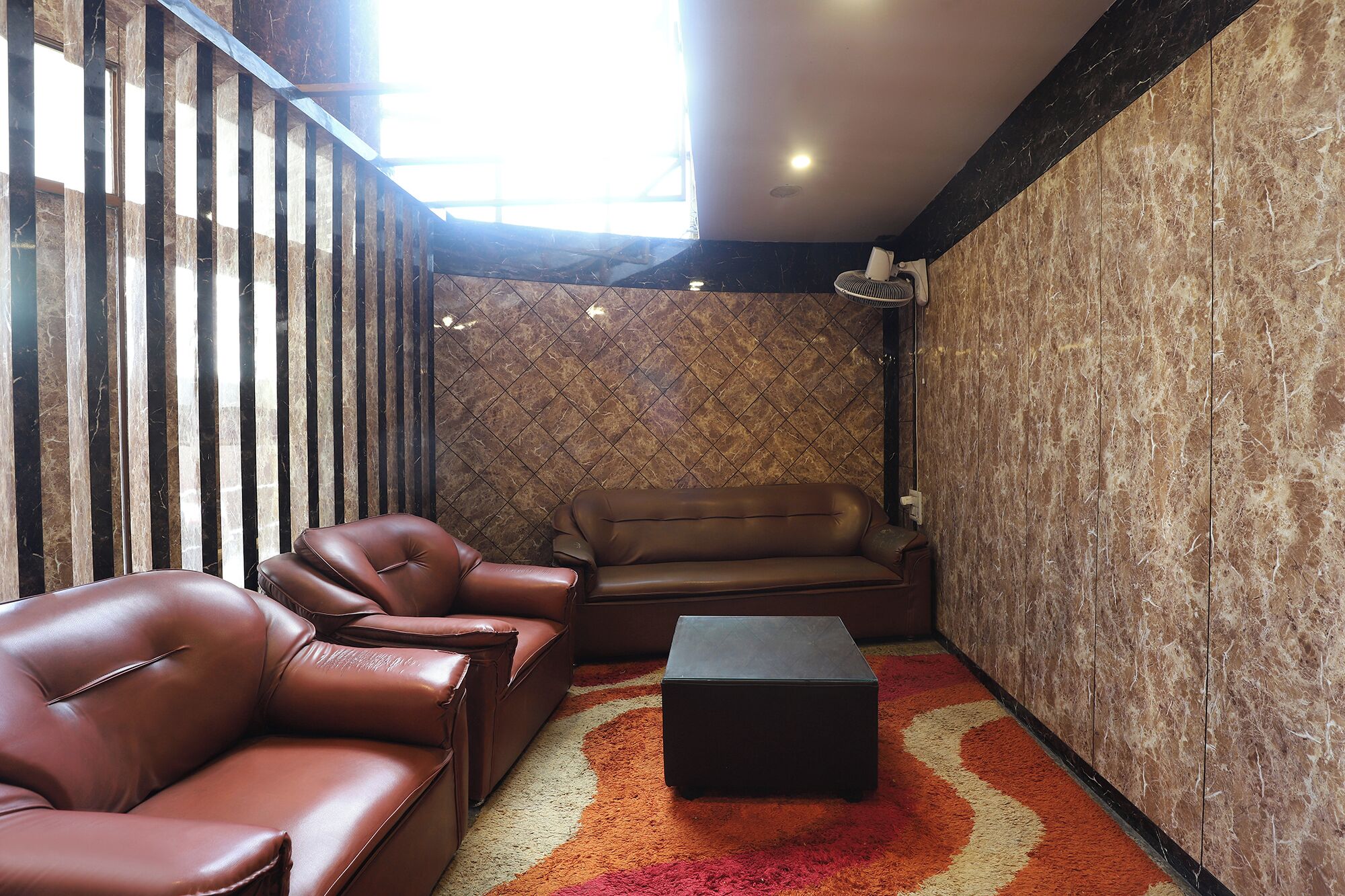 A corner room with a brown leather sofa, a matching armchair, and a black ottoman on a patterned carpet.