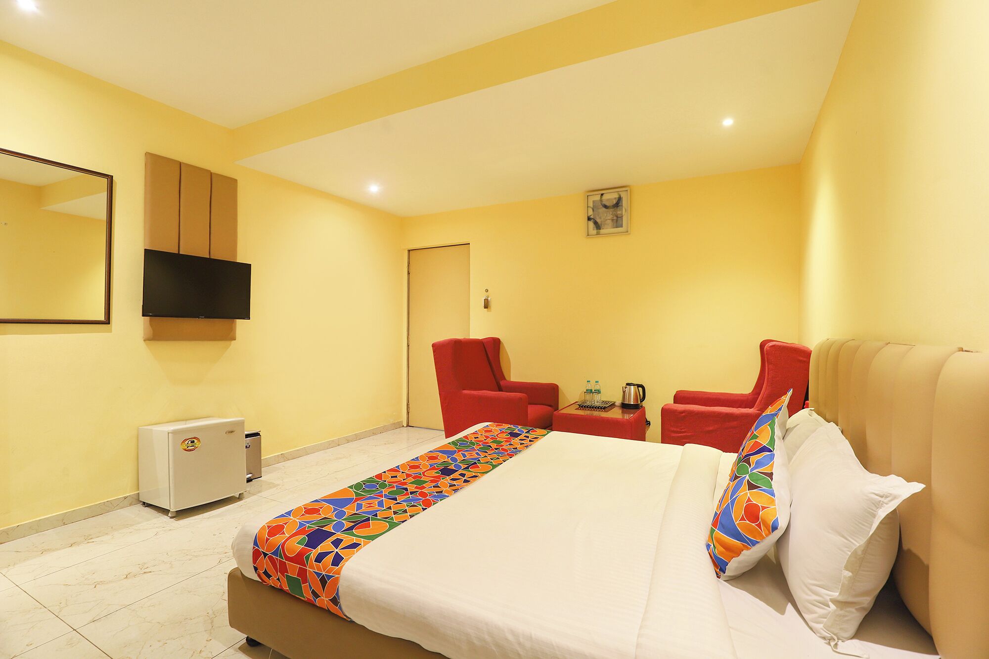 A hotel room with a bed, two red armchairs, a small table with a kettle, a TV, and a minibar.