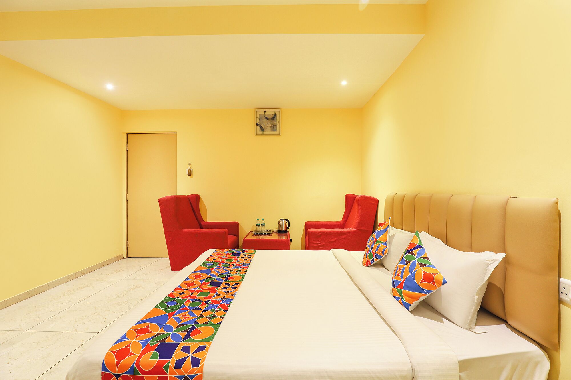 A hotel room with a bed, two red armchairs, a small table with a kettle, and a colorful runner.