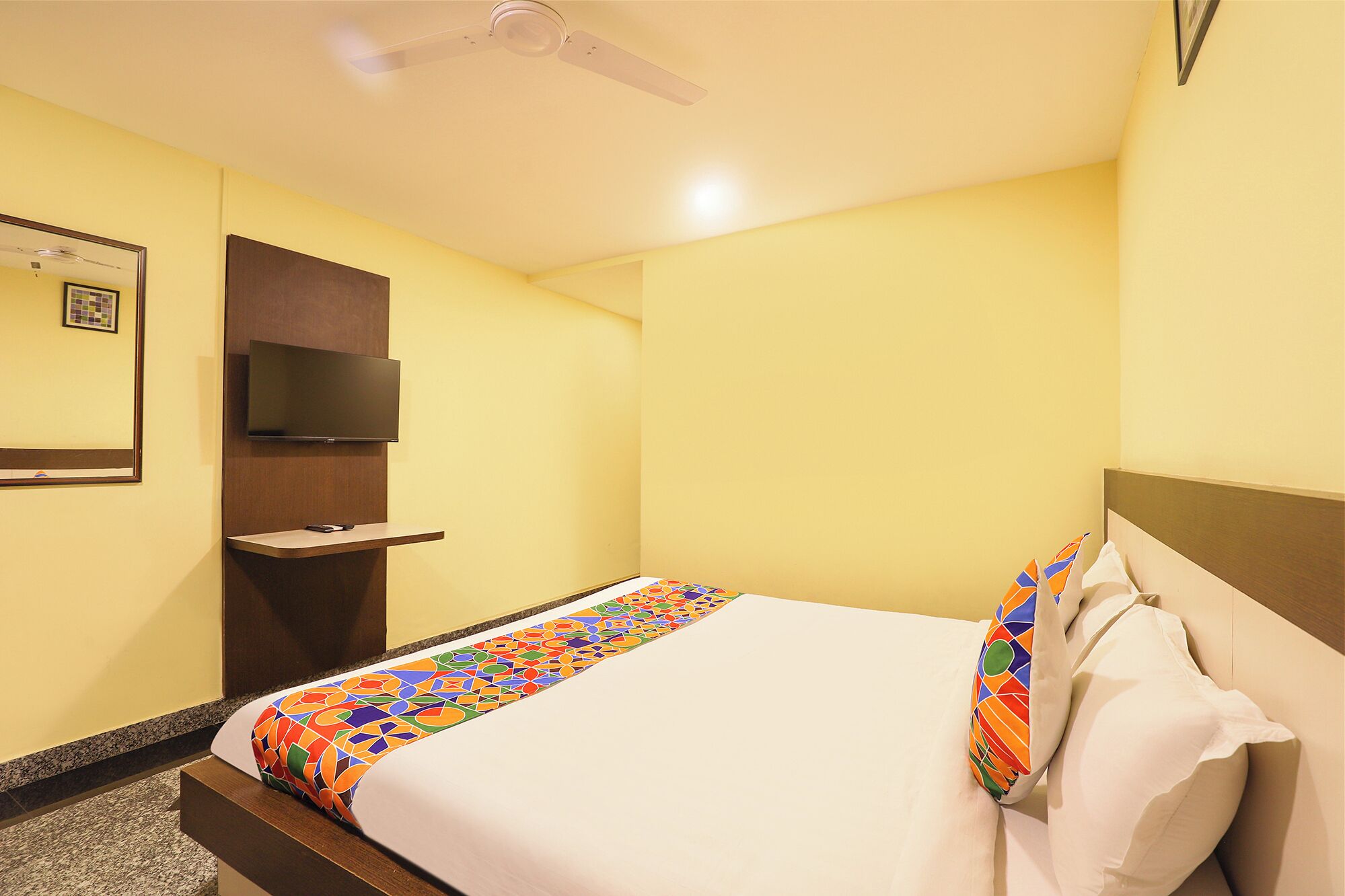 A hotel room with a bed, a TV mounted on the wall, a ceiling fan, and a small shelf.