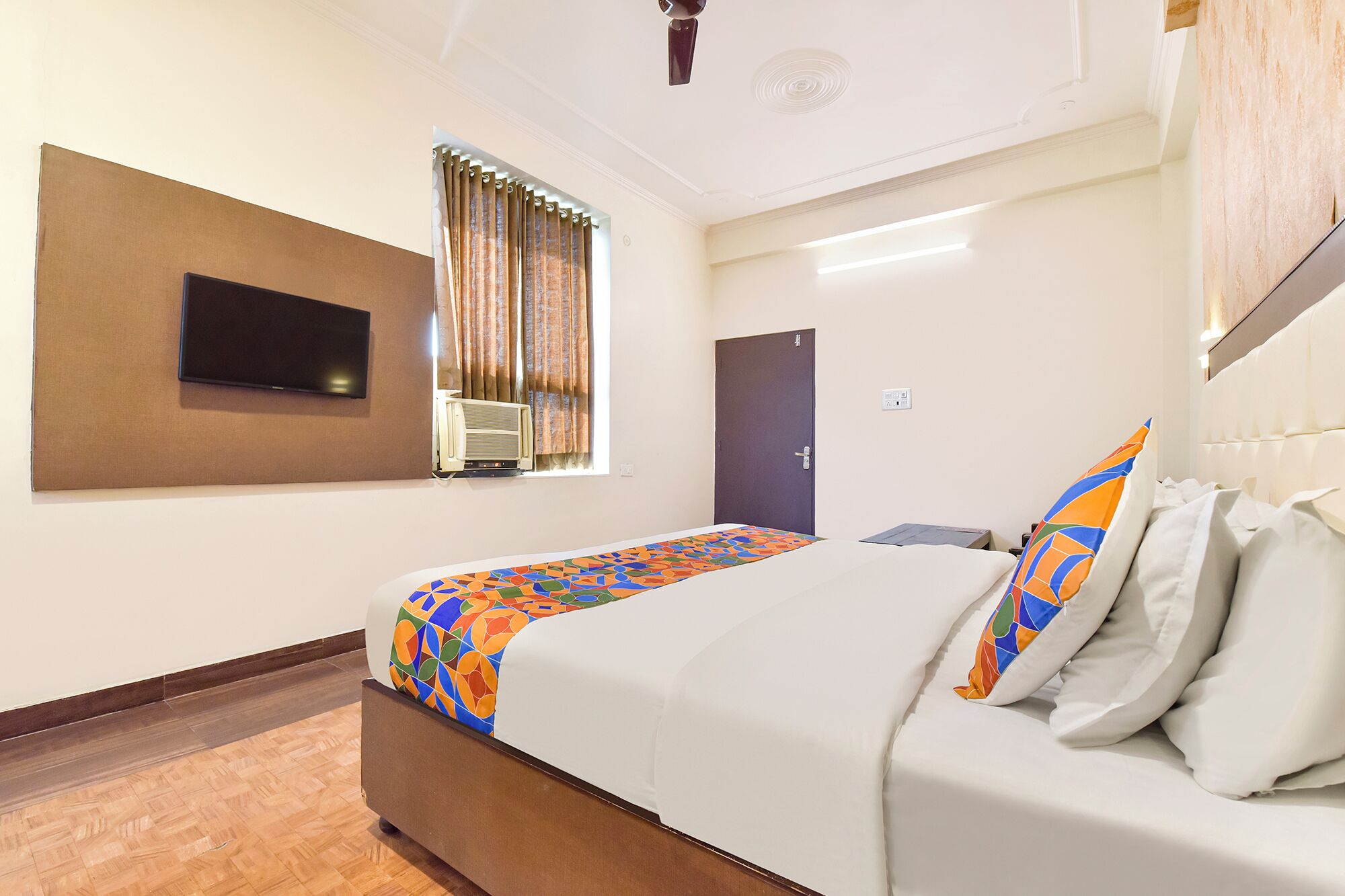 A hotel room with a bed, a television mounted on the wall, an air conditioning unit, a door, and a window with curtains.