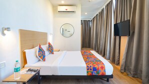 Una camera d'albergo con un letto, un comodino con una bottiglia e telecomandi, uno specchio, un condizionatore d'aria, una televisione e una tenda.