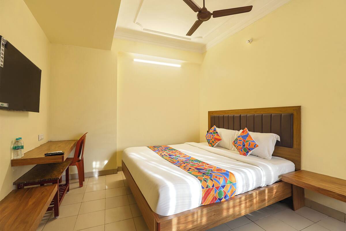 Une chambre d’hôtel avec un lit, une tête de lit en bois, un bureau avec une chaise, un téléviseur fixé au mur et un ventilateur de plafond.