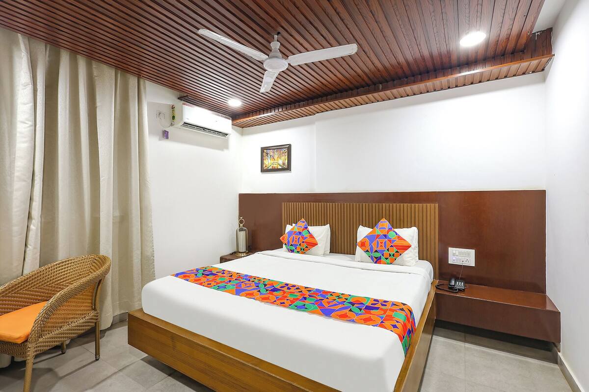 A hotel room with a wooden ceiling, a bed with colorful pillows, a wicker chair, and a bedside table with a phone.