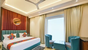 Una habitación de hotel moderna con una cama grande, dos sillones, una mesita, un ventilador de techo y un amplio ventanal con cortinas.