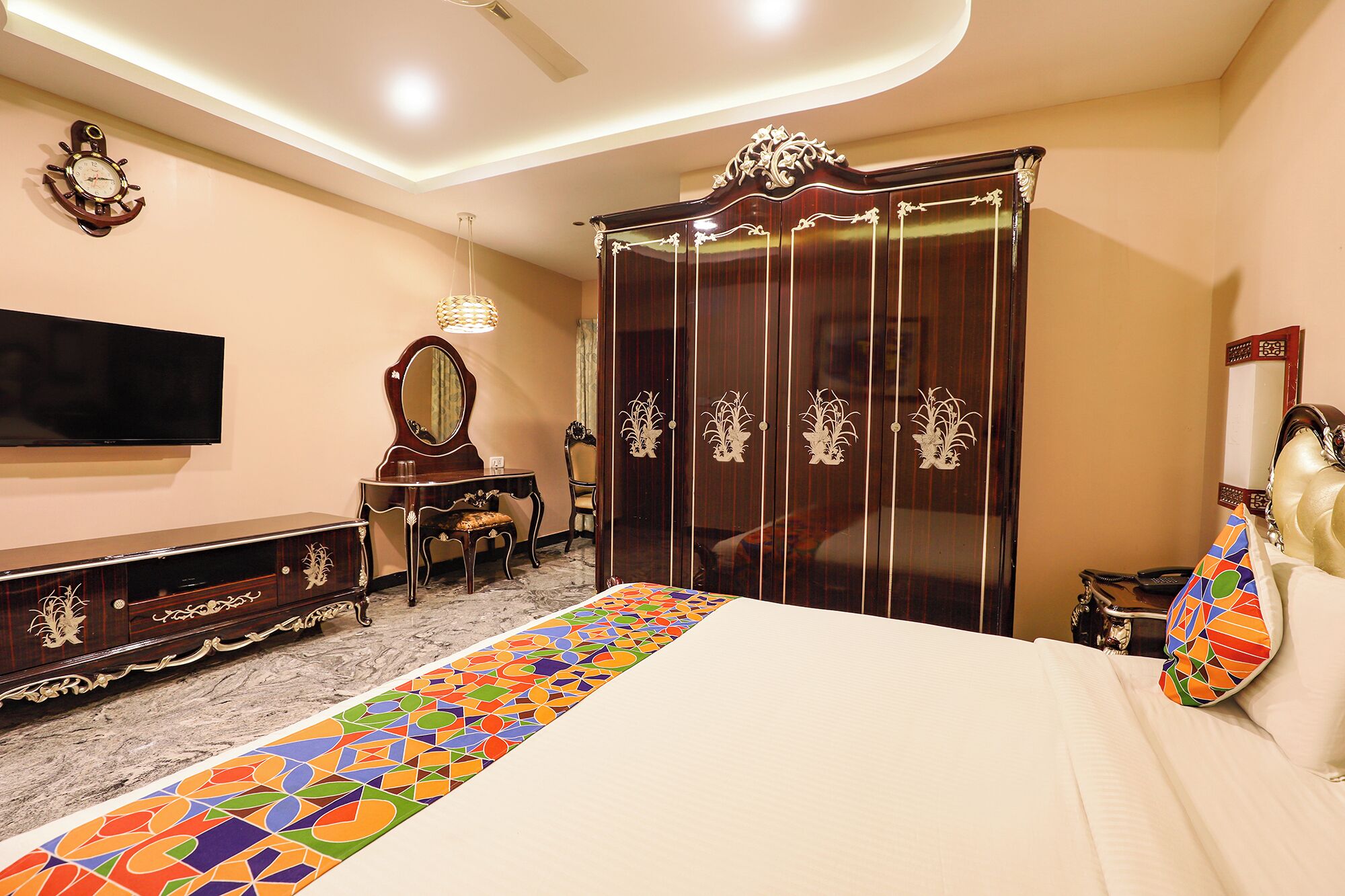 Quarto de hotel com uma cama, televisão, guarda-roupa, penteadeira com espelho e tapete estampado.