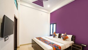 A modern hotel room with a large bed, a TV mounted on the wall, and a purple accent wall. - FabExpress Awadh Royal (Faizabad)