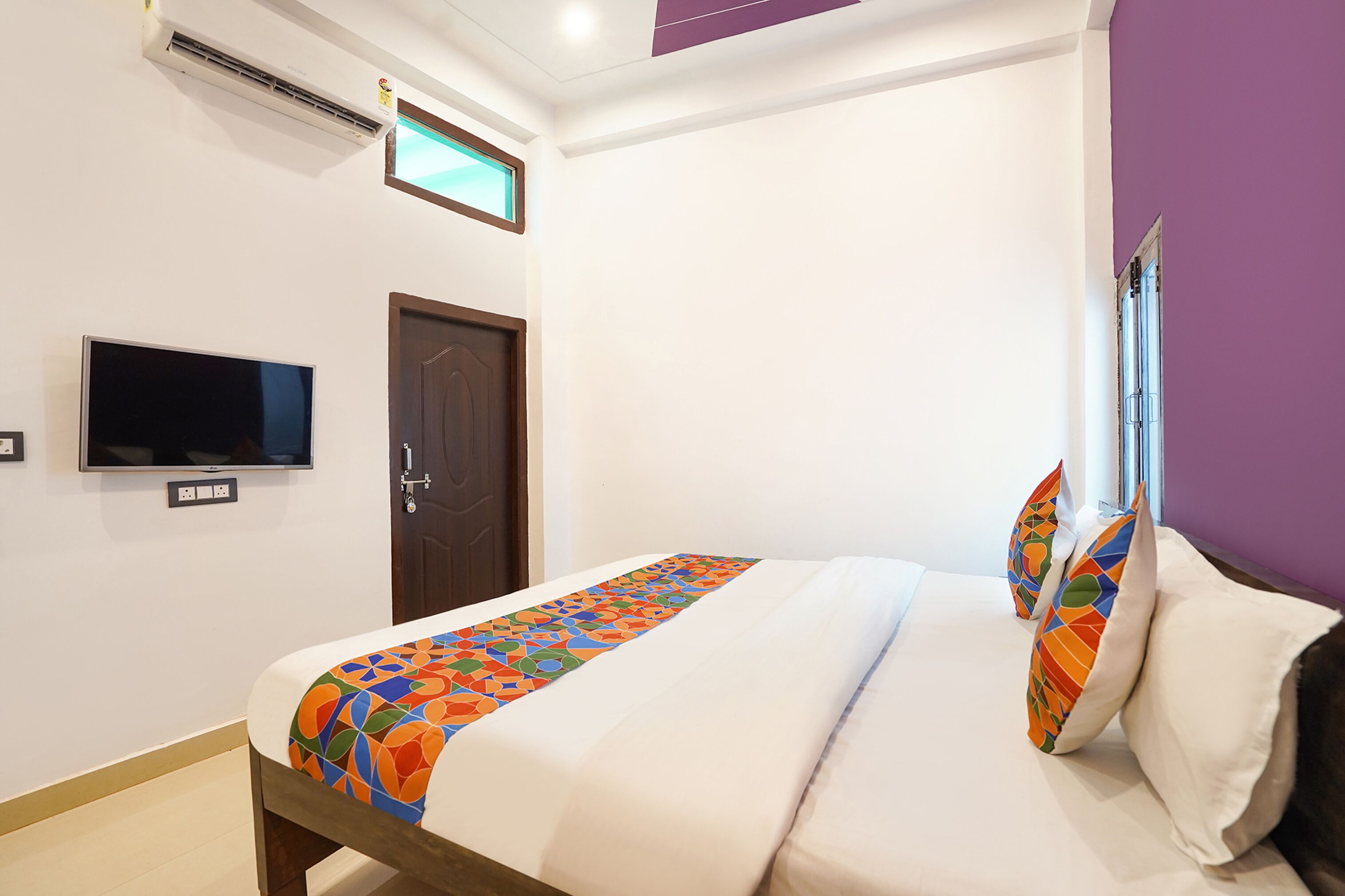 A hotel room with a bed, a TV, an air conditioning unit, and a door.