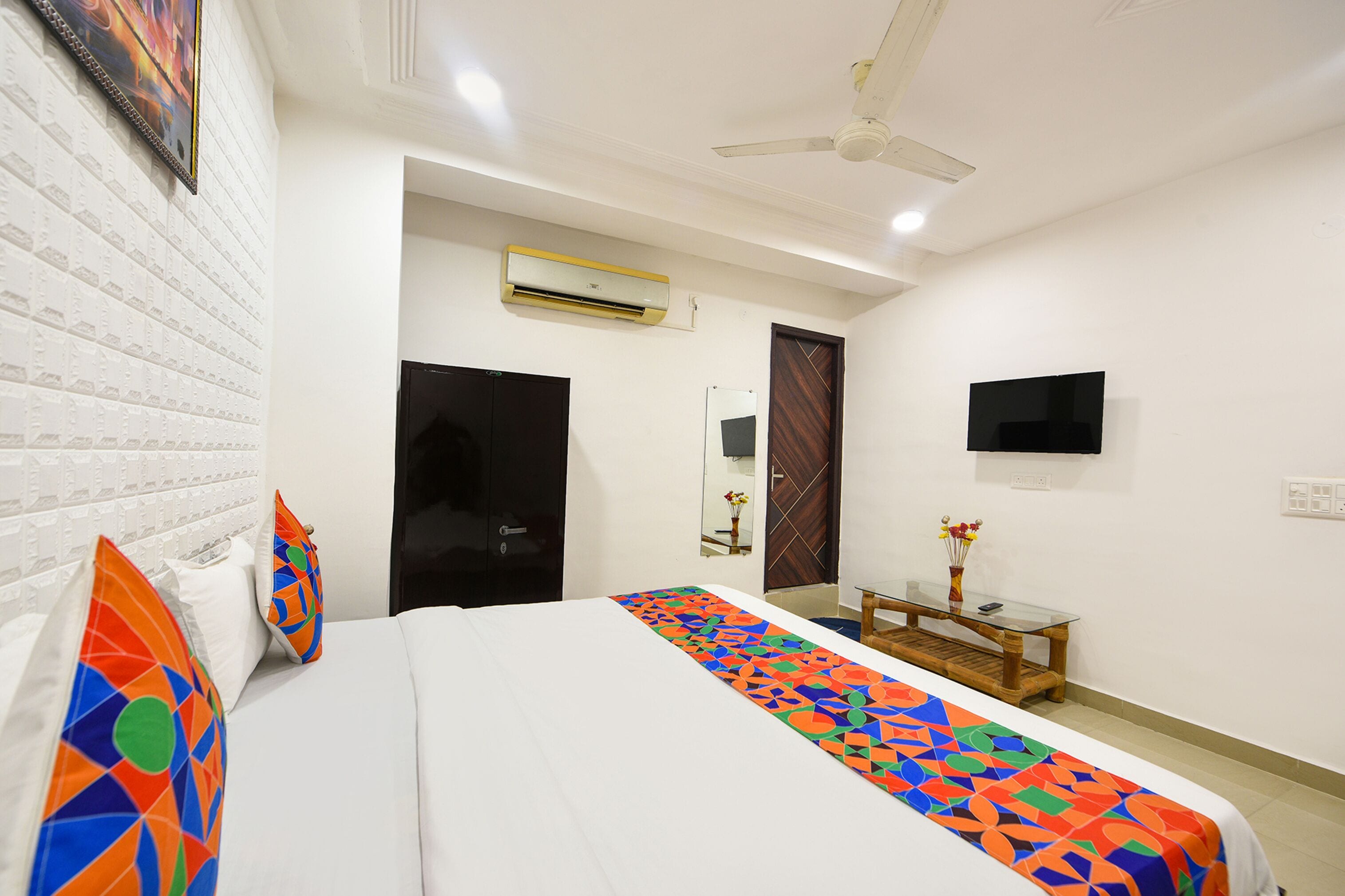 A hotel room with a bed, a ceiling fan, an air conditioning unit, a TV, and a small table with a vase.