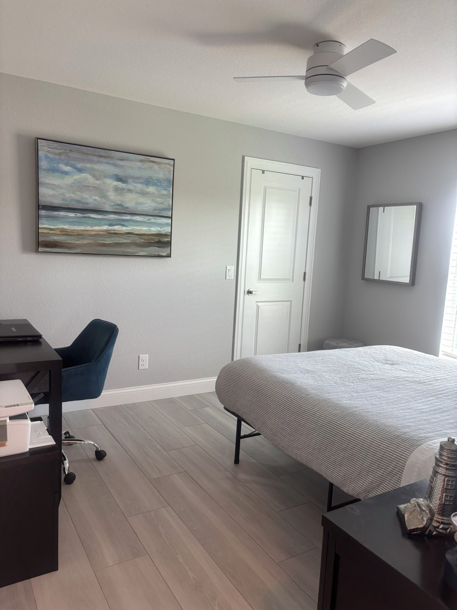 A bedroom with a bed, a desk with a chair, a ceiling fan, a door, and a painting on the wall.