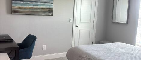 A bedroom with a bed, a desk with a chair, a ceiling fan, a door, and a painting on the wall.
