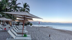 Una playa con sillas de descanso blancas y sombrillas, palmeras y vistas al océano y montañas.