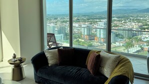 Una sala de estar moderna con un sofá azul oscuro, una mesa de centro de vidrio y vistas de un paisaje urbano a través de grandes ventanales.