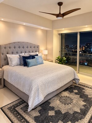 Un dormitorio con una cama grande, un ventilador de techo, una mesita de noche, una lámpara y vistas a un paisaje urbano a través de un ventanal.