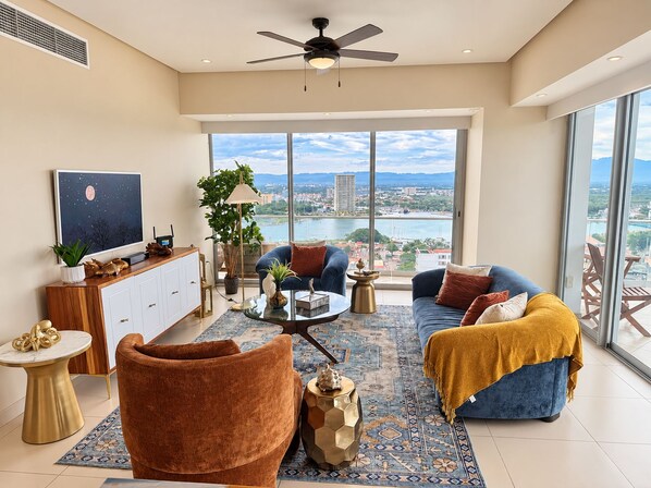 Un amplio salón con un ventanal grande, un ventilador de techo y vistas a un paisaje urbano.