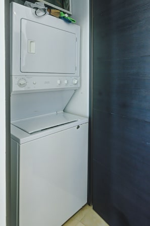 A compact, white laundry room with a built-in washer and dryer, a small shelf with cleaning supplies, and a door with a textured pattern.