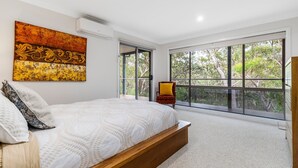 A bedroom with a large bed, a chair, and a view of trees through large windows.