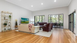 A spacious living room with a white sofa, a brown leather chair, and a coffee table. There is a wooden floor, a white wall, and a large window with a view of trees.