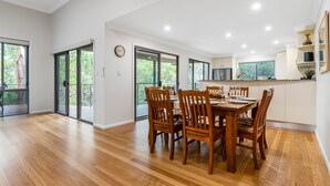 A spacious dining area with a wooden table and chairs, large windows, and a view of greenery outside.