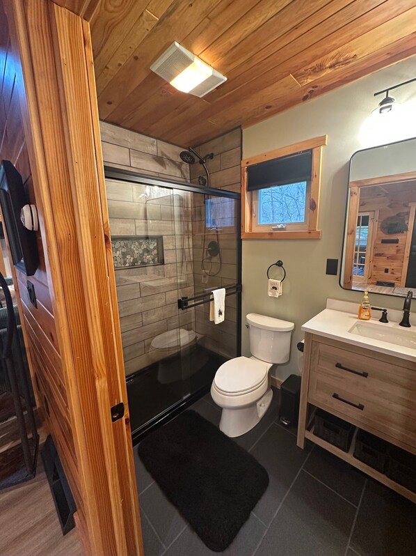 A compact bathroom with a glass-enclosed shower, a white toilet, and a wooden vanity with a mirror. - Charming, NEWLY Renovated, 3 Bedroom Cabin by the Creek, Minutes from 7 Springs (Champion)
