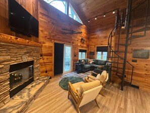 A cozy log cabin interior with a stone fireplace, a large sectional sofa, a wooden chair, and a spiral staircase. - Charming, NEWLY Renovated, 3 Bedroom Cabin by the Creek, Minutes from 7 Springs (Champion)
