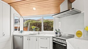 A modern kitchen with white cabinetry, a stainless steel dishwasher, a double oven, and a large window offering a view of a garden and hills. - Arrowtown Family Home with spa (Arrowtown)