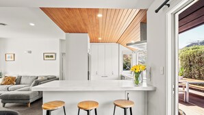 Une cuisine moderne avec un plan de travail blanc, des tabourets en bois et une vue sur l’extérieur.