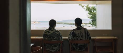 Deux personnes sont assises à une table en bois près d’une fenêtre, d’où l’on peut admirer une plage et des montagnes.