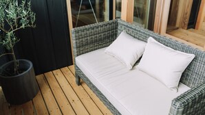 Un sofá de mimbre con cojines blancos sobre una terraza de madera, una planta en maceta y una puerta de vidrio al fondo.
