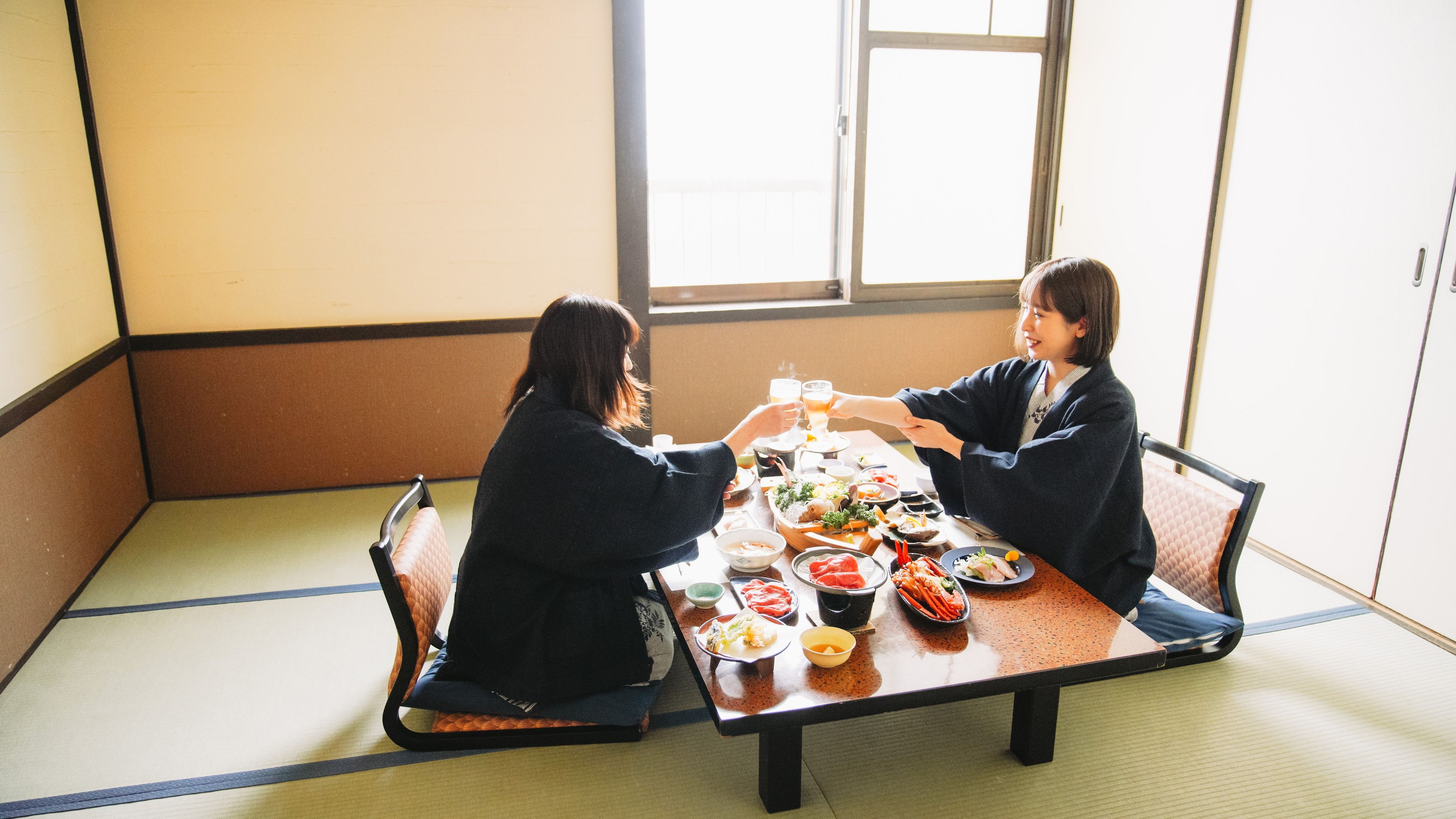 Dos personas sentadas en una mesa japonesa tradicional con varios platos, en una habitación con suelo de tatami y una ventana.