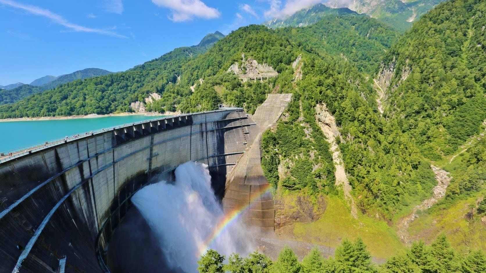 A dam with a waterfall and a rainbow, surrounded by lush green mountains and a clear blue sky.