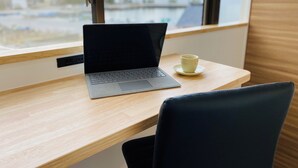 Una computadora portátil sobre un escritorio de madera, con una taza en un platillo, cerca de una ventana con vistas a edificios.