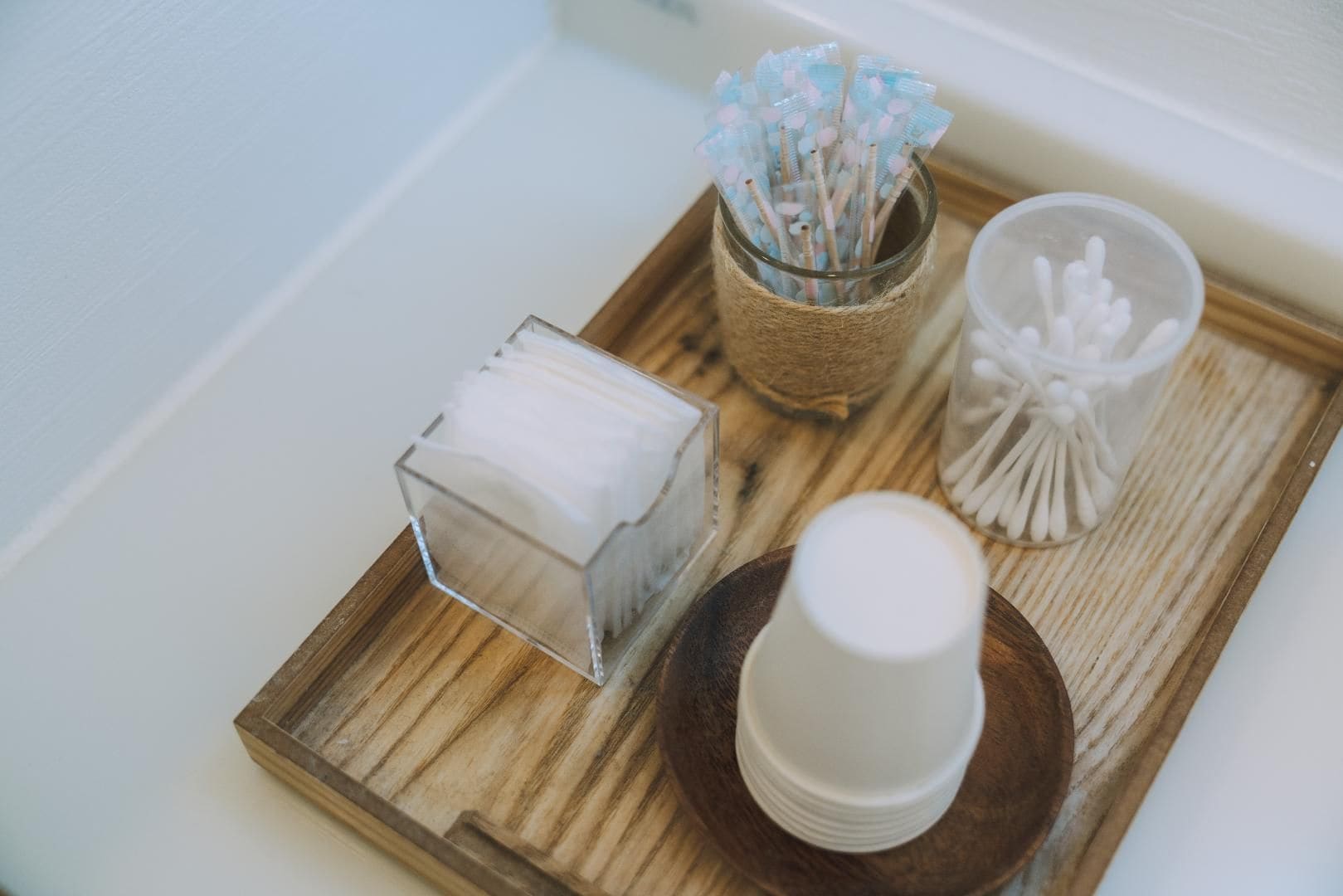 Una bandeja de madera con un recipiente de vidrio lleno de bastoncillos de algodón, un tarro con bastoncillos de algodón y una pila de vasos blancos.