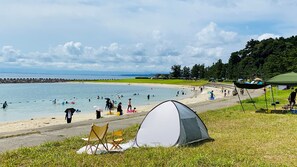 Una playa con una tienda de campaña y sillas dispuestas sobre el césped, un rompeolas rocoso al fondo, y gente nadando y caminando por la orilla.