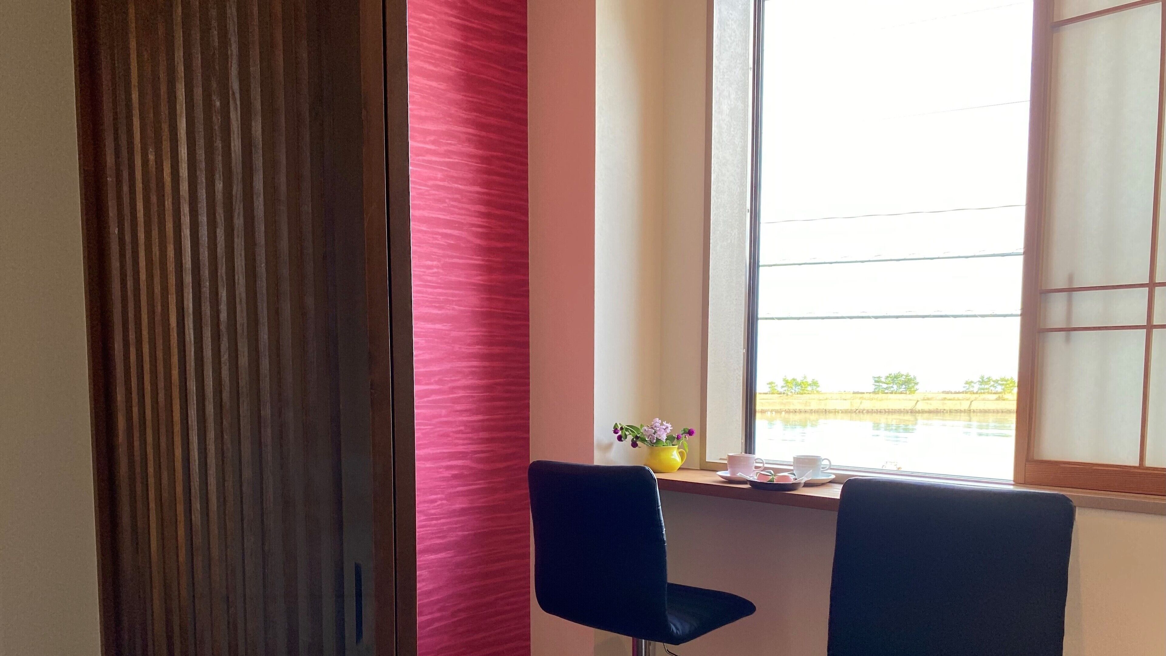 Una habitación con puerta de madera, pared rosa, ventana con vistas al agua y dos sillas.