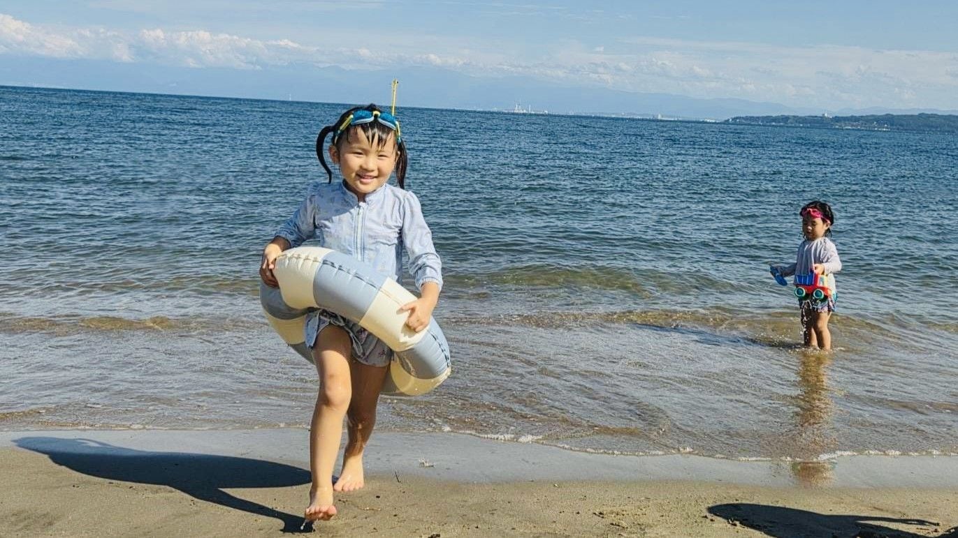 A beach scene with a child holding a beach toy and another child standing in the water.