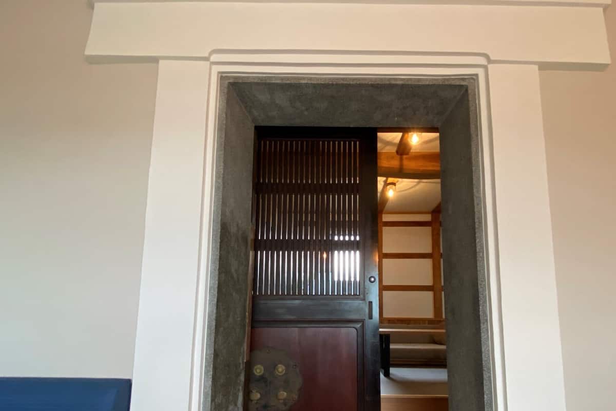 Un acceso con una puerta de madera oscura y una celosía metálica, que da a una escalera de madera y una pared blanca.