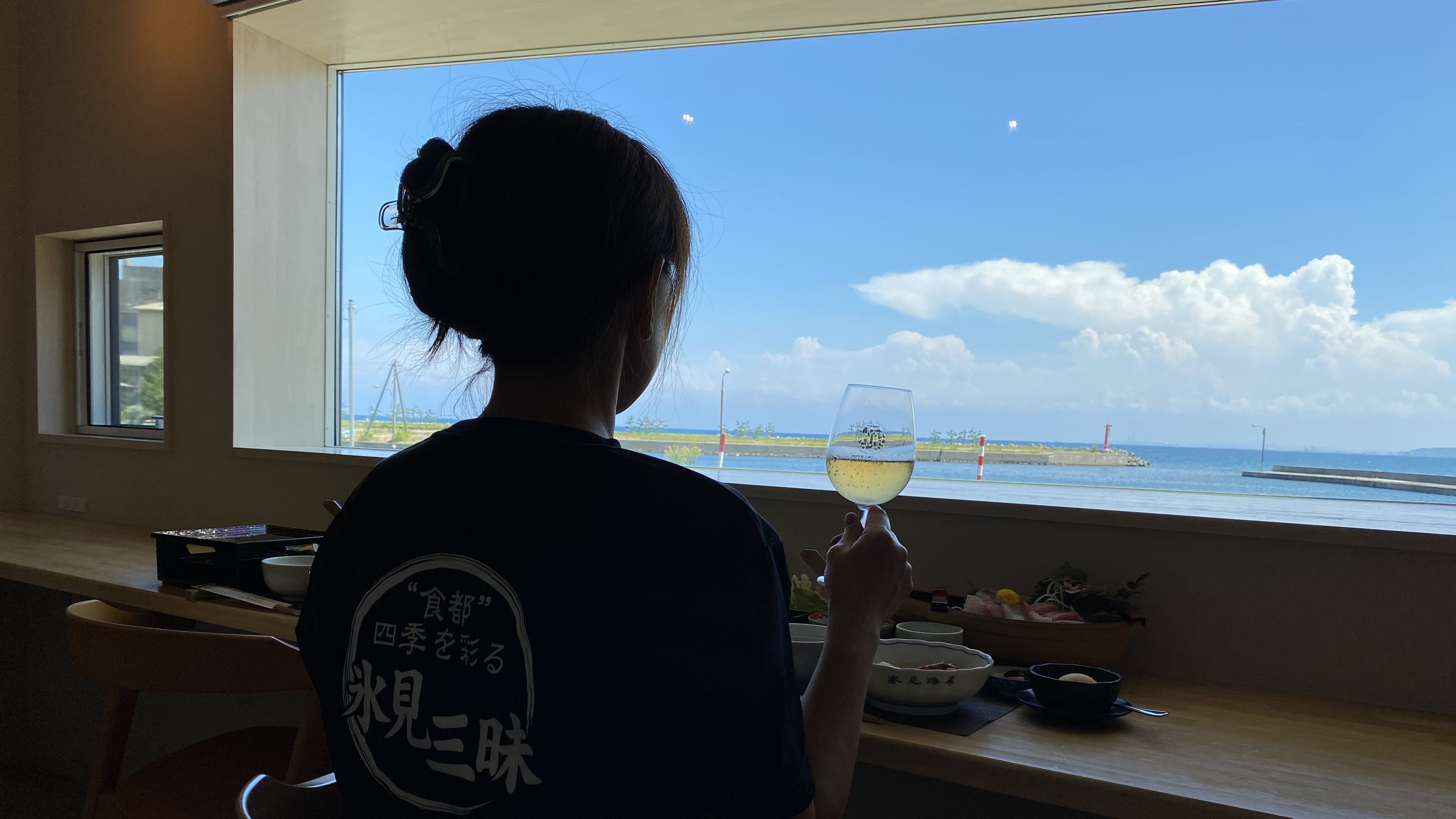 A person seated by a window with a view of the sea, holding a glass of white wine.