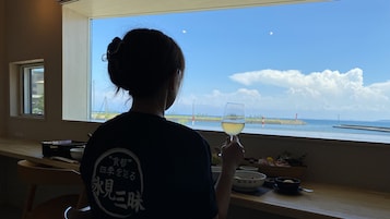 Una persona sentada junto a una ventana con vista al mar, sosteniendo una copa de vino blanco.