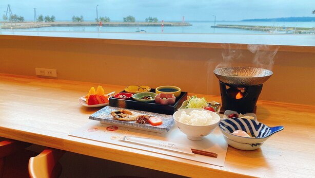 Mesa con varios platos y un fogón portátil con vapor, vista desde una ventana con vista al agua.