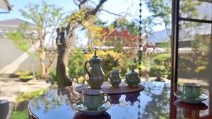 A round table with a tea set including a teapot, cups, and saucers, set outdoors with a view of trees and a building in the background. - Kashibesso Sakuraso (Beppu)