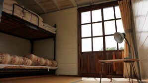 A dormitory room with bunk beds, a wooden door, a window with curtains, and a small table with a lamp. - Kuju Aid Station (Kokonoe)