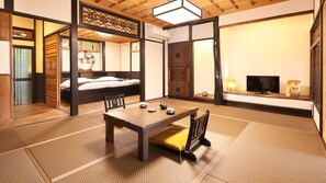 Una habitación tradicional japonesa con suelo de tatami, una mesa baja con juego de té, una cama con cabecero de madera y un televisor sobre una estantería.